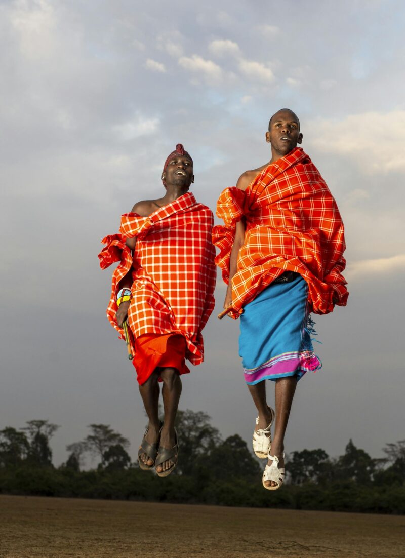 Two Maasai men in traditional attire mid-jump outdoors in Kenya, showcasing cultural heritage.